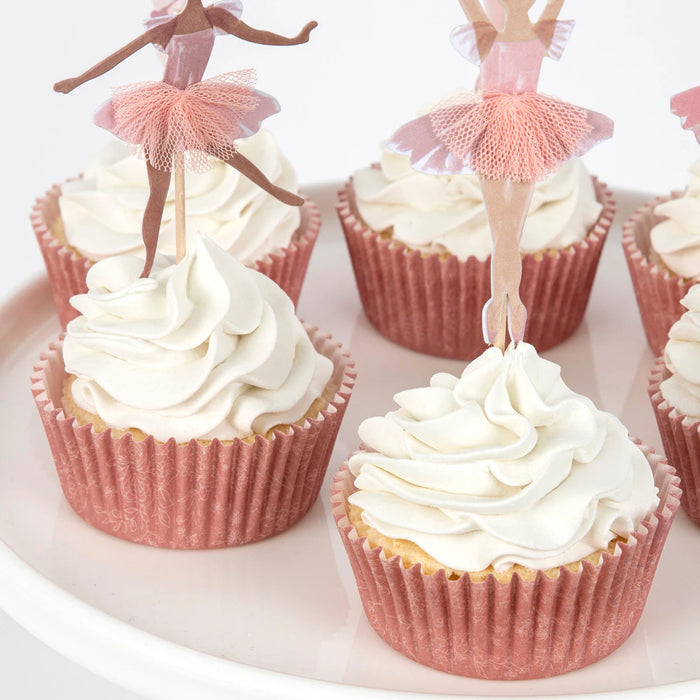 Cupcakes with white frosting and pink cupcake liners, topped with ballerina figurines on a white plate.