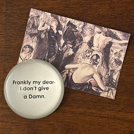 Glass paperweight featuring the iconic quote “Frankly my dear, I don’t give a damn” in black typewriter-style text, placed on a wooden surface beside a vintage-style art postcard of a glamorous ballroom scene.