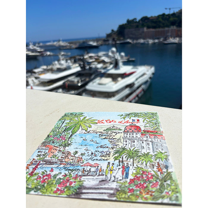 Colorful illustration of a coastal scene with palm trees and buildings on a napkin placed on a ledge with a marina in the background.