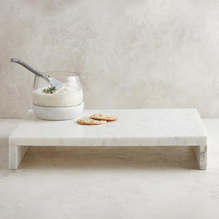 A white marble waterfall stand with a glass bowl and silver serving utensil on top, accompanied by two crackers.