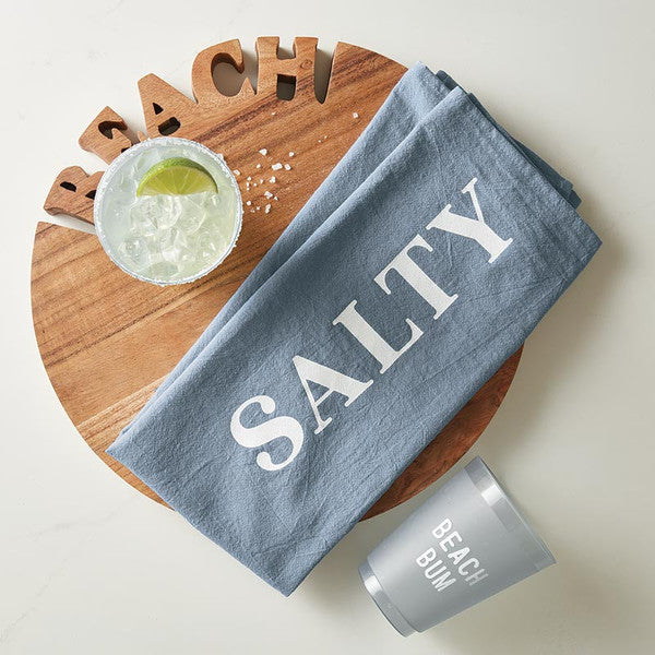 Blue towel with 'Salty' text on a wooden board with a margarita and 'Beach Bum' cup. Wooden round sign with 'BEACH' cut out of the top. Handcrafted acacia wood tray with ‘BEACH’ lettering, styled with summer appetizers and coastal table accessories.