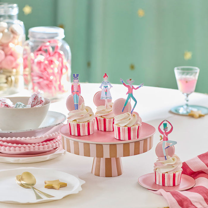 Cupcakes with decorative toppers on a pink striped cake stand with candy and plates in the background.
