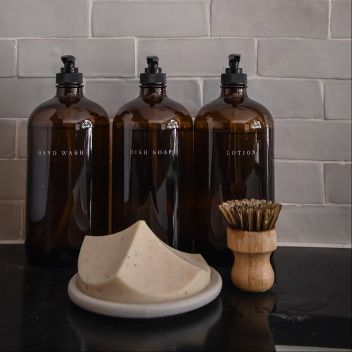 Three brown bottles with black caps, a white soap dish, and a wooden brush on a dark surface against a gray tiled wall.