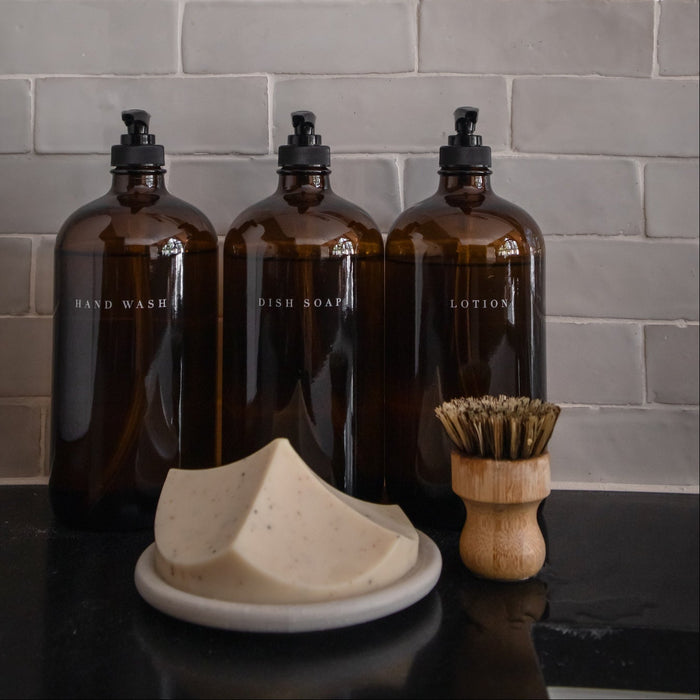 Three brown bottles with black caps, a white soap dish, and a wooden brush on a dark surface against a gray tiled wall.