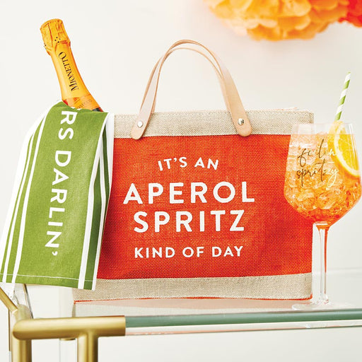 Tote bag with Aperol Spritz text, bottle of Prosecco, and cocktail on a table.