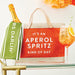 Tote bag with Aperol Spritz text, bottle of Prosecco, and cocktail on a table.