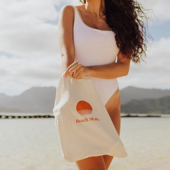 Woman holding a white tote bag with 'Beach Slats' logo on a beach.