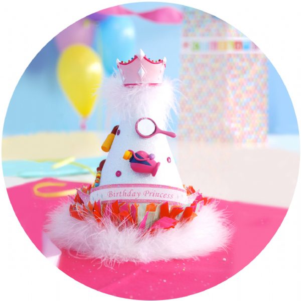 A colorful birthday party hat with white and pink glitter, marabou feathers, and a jeweled crown topper, displayed against a festive background with balloons.