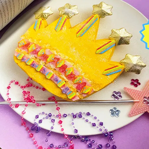 A yellow crown-shaped cake decorated with gold star toppers, rainbow stripes, and rows of colorful candy,.