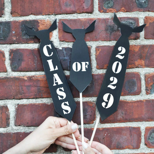 Three black paper tie photo props on wooden dowels spelling out "Class of 2029" held against a red brick wall — printable graduation party photo booth props