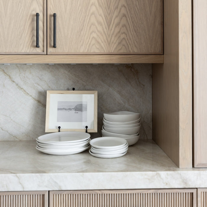Stacks of Drift Appetizer Plates in Matte White on a kitchen shelf with wooden cabinets and marble backsplash.