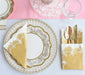 Elegant table setting with gold and white paper plates, napkins, and cutlery on a pink tablecloth.