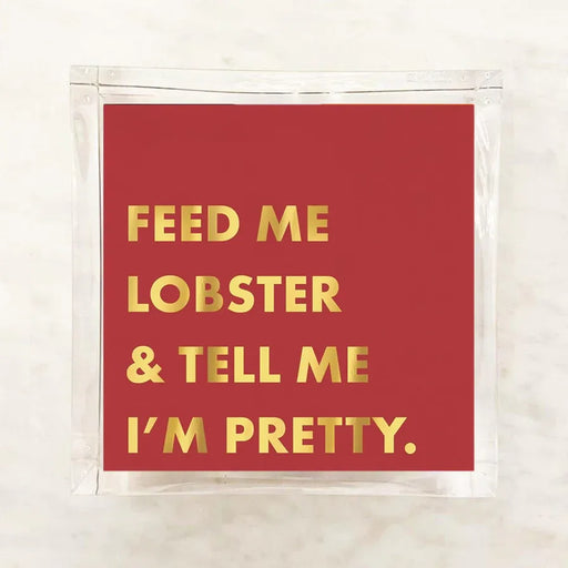 A square red card or sticker with gold foil text reading 'FEED ME LOBSTER & TELL ME I'M PRETTY.' inside a clear plastic sleeve, placed on a light marble-like surface.