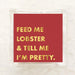 A square red card or sticker with gold foil text reading 'FEED ME LOBSTER & TELL ME I'M PRETTY.' inside a clear plastic sleeve, placed on a light marble-like surface.