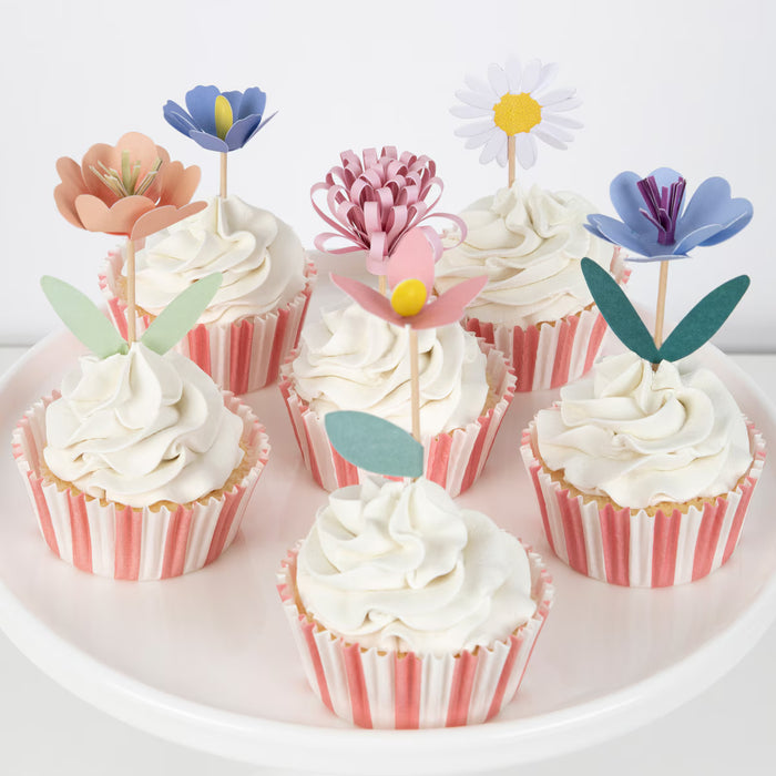 Cupcakes with white frosting and pink striped wrappers, decorated with colorful flower toppers on a white background.