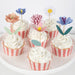 Cupcakes with white frosting and pink striped wrappers, decorated with colorful flower toppers on a white background.
