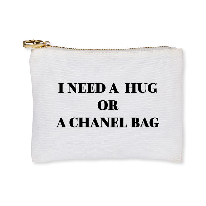 A white canvas zipper pouch with the text 'I NEED A HUG OR A PARIS BAG' printed in black. It features a gold metal zipper with a metal pull.