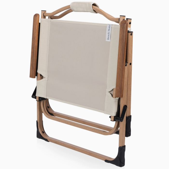 A folding outdoor lounge chair with a beige canvas seat and backrest, supported by a light brown wooden frame with black plastic feet, shown on a white background.