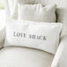 White lumbar pillowcase with the words “Love Shack” in black lettering styled on a neutral chair