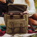 Green backpack with brown straps held by a person sitting on a colorful blanket outdoors.