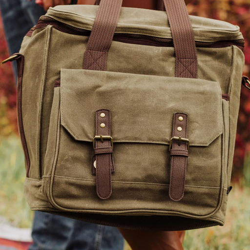 Green canvas bag with brown straps and buckles held by a person in an outdoor setting.