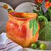 Decorative ceramic peach pitcher with fruit design on a table with limes and a plant.