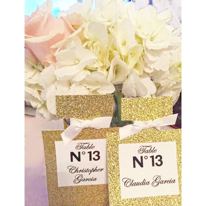 A gold glittered perfume bottle with a custom label displaying a table number and name, set against a floral backdrop.