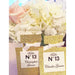 A gold glittered perfume bottle with a custom label displaying a table number and name, set against a floral backdrop.