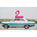 Vintage car with a pink flamingo float on top, driving on a road with a scenic background.