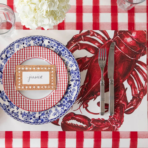 Place setting with lobster-themed placemat, checkered plates, and cutlery on a red and white checkered tablecloth.