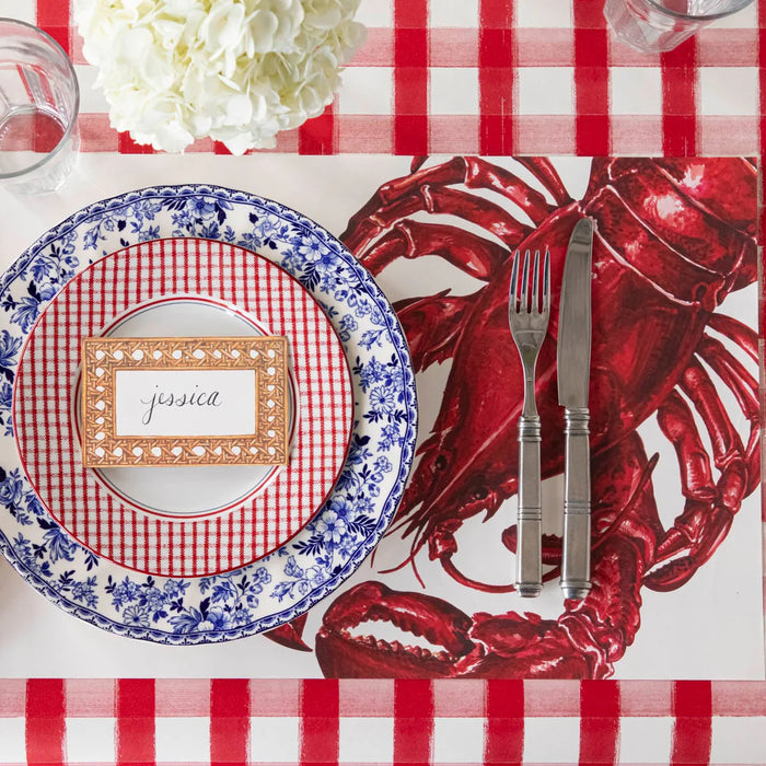 Place setting with lobster-themed placemat, checkered plates, and cutlery on a red and white checkered tablecloth.