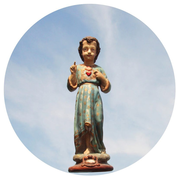A statue of the Sacred Heart Child with a blue robe, holding a heart, with gold details and a sky background.