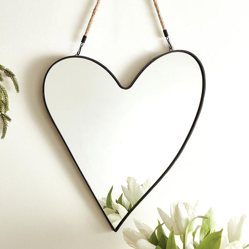 Heart-shaped mirror with black frame hanging on a white wall with plants below.