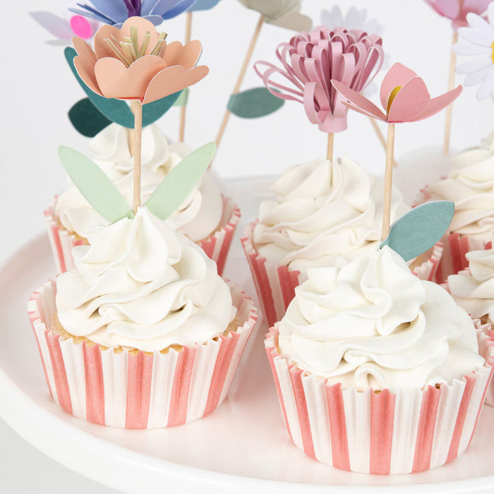 Cupcakes with white frosting and pink striped wrappers, topped with floral picks on a white plate.