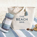 Beige tote bag with 'THE BEACH BAG' text on a striped towel with a beach towel and sandals.