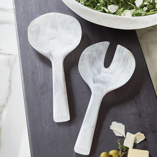 Two White Resin salad servers on a dark surface with a bowl of salad in the background.