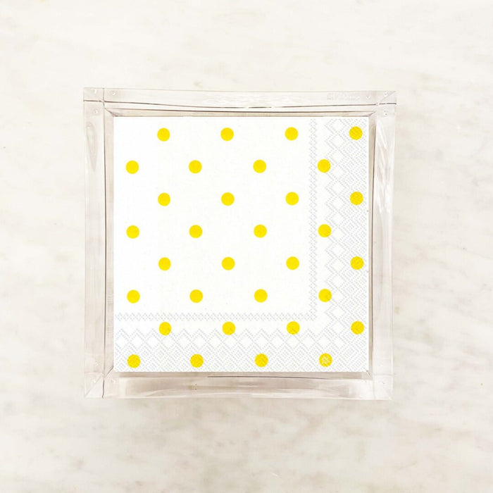 A clear napkin holder box containing yellow and white polka dot cocktail napkins displayed on a marble background.