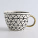 Ceramic mug with geometric black pattern and gold handle on a light gray background