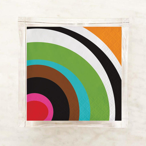 A square paper napkin with a retro concentric circle pattern in green, turquoise, brown, black, white, orange, and pink, sealed in a clear plastic sleeve on a light marble surface.