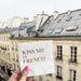 White cocktail napkin with gold foil Kiss Me in French lettering against a Paris rooftop view