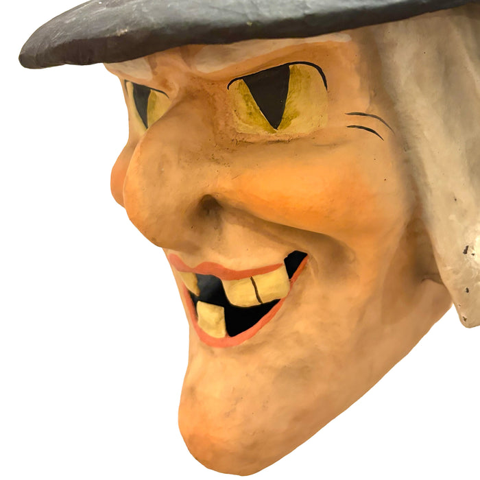 A close-up of a papier-mâché witch bucket with a wide grin, yellow eyes, and a black which hat.