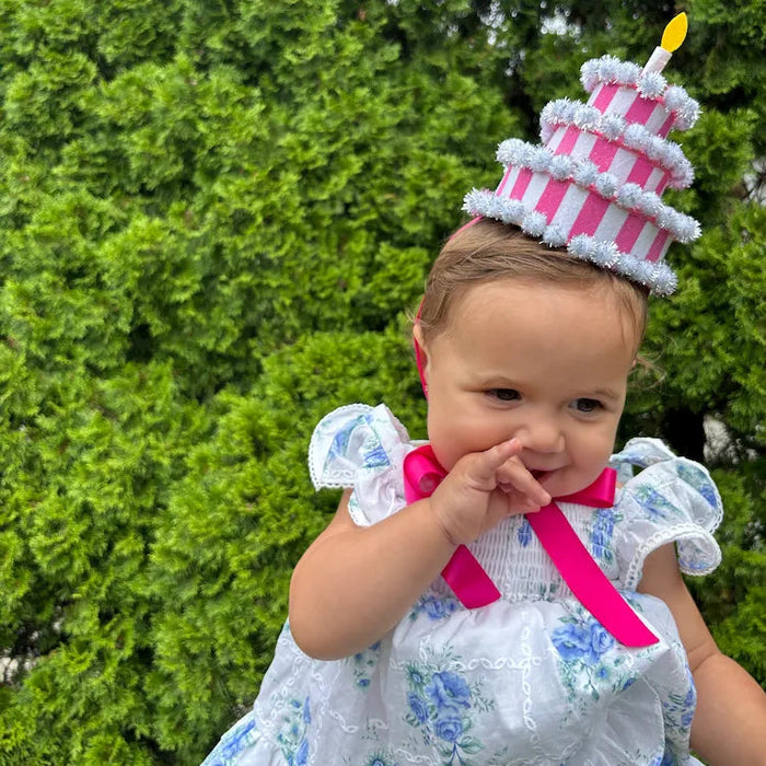 A toddler wearing a pink and white striped tiered cake hat with a yellow candle.