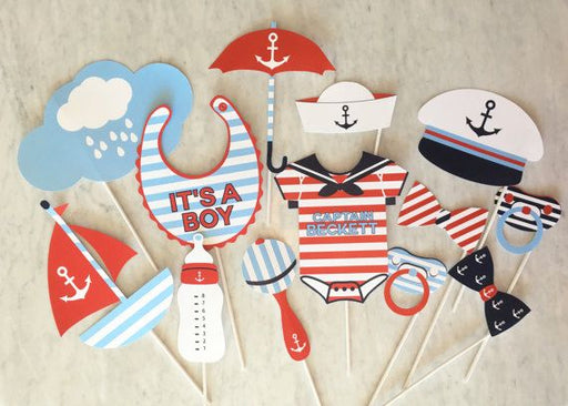 Set of 12 red white and blue nautical baby shower photo props including a personalized bib, onesie, sailboat, captain's hat, pacifier, rattle, and striped bow ties