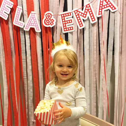 Retro red and white printable name banner spelling "ELA & EMA" hanging over a streamer backdrop with a young girl holding popcorn below