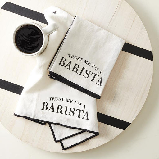 Two white towels with 'Trust Me I'm a Barista' text on a wooden surface with a cup of coffee.