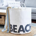Large white canvas beach storage tote bag with bold BEACH lettering and waterproof laminated lining — lifestyle shot in coastal living room